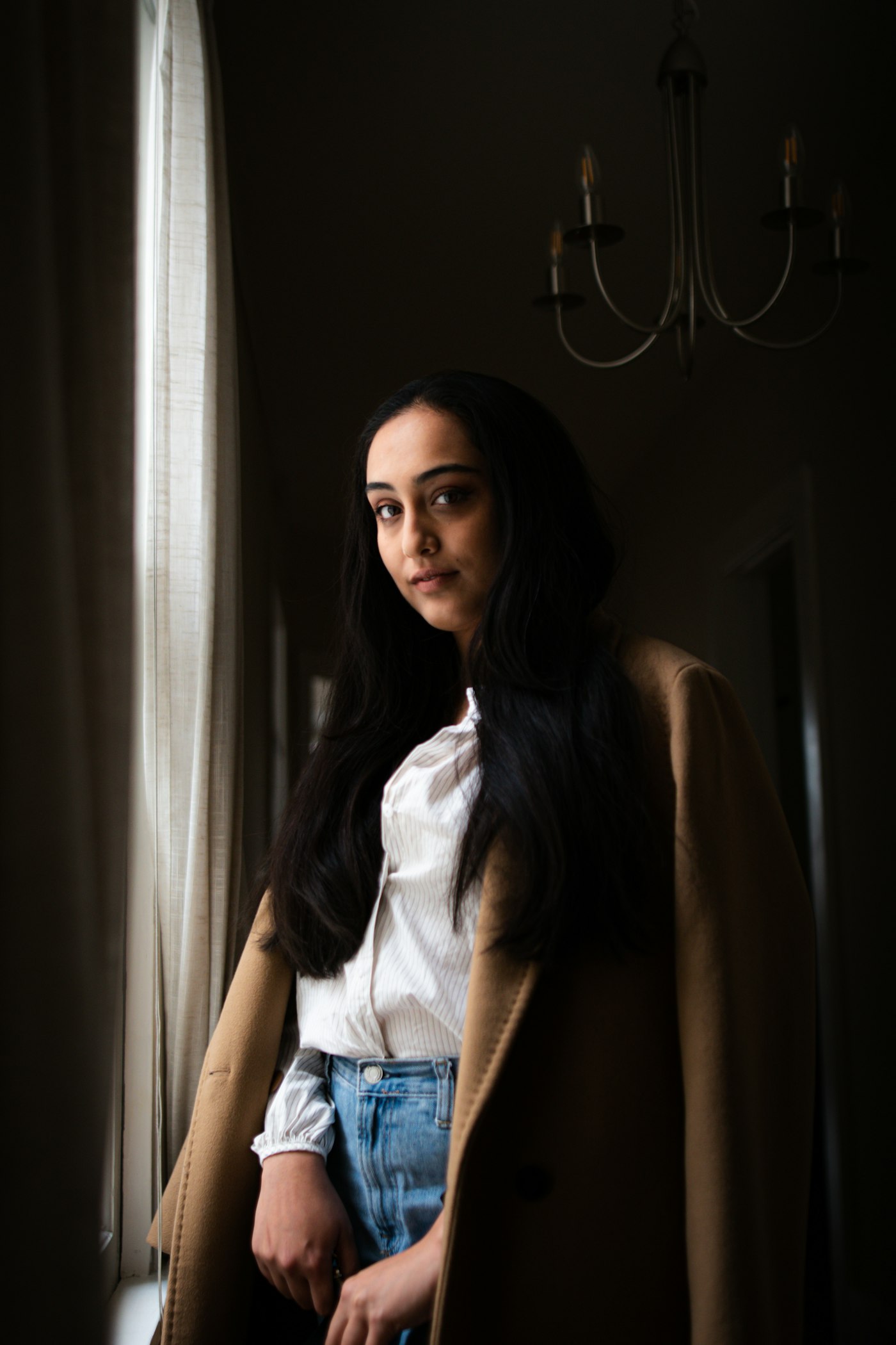 A woman in a camel coat standing in window light in a warm interior