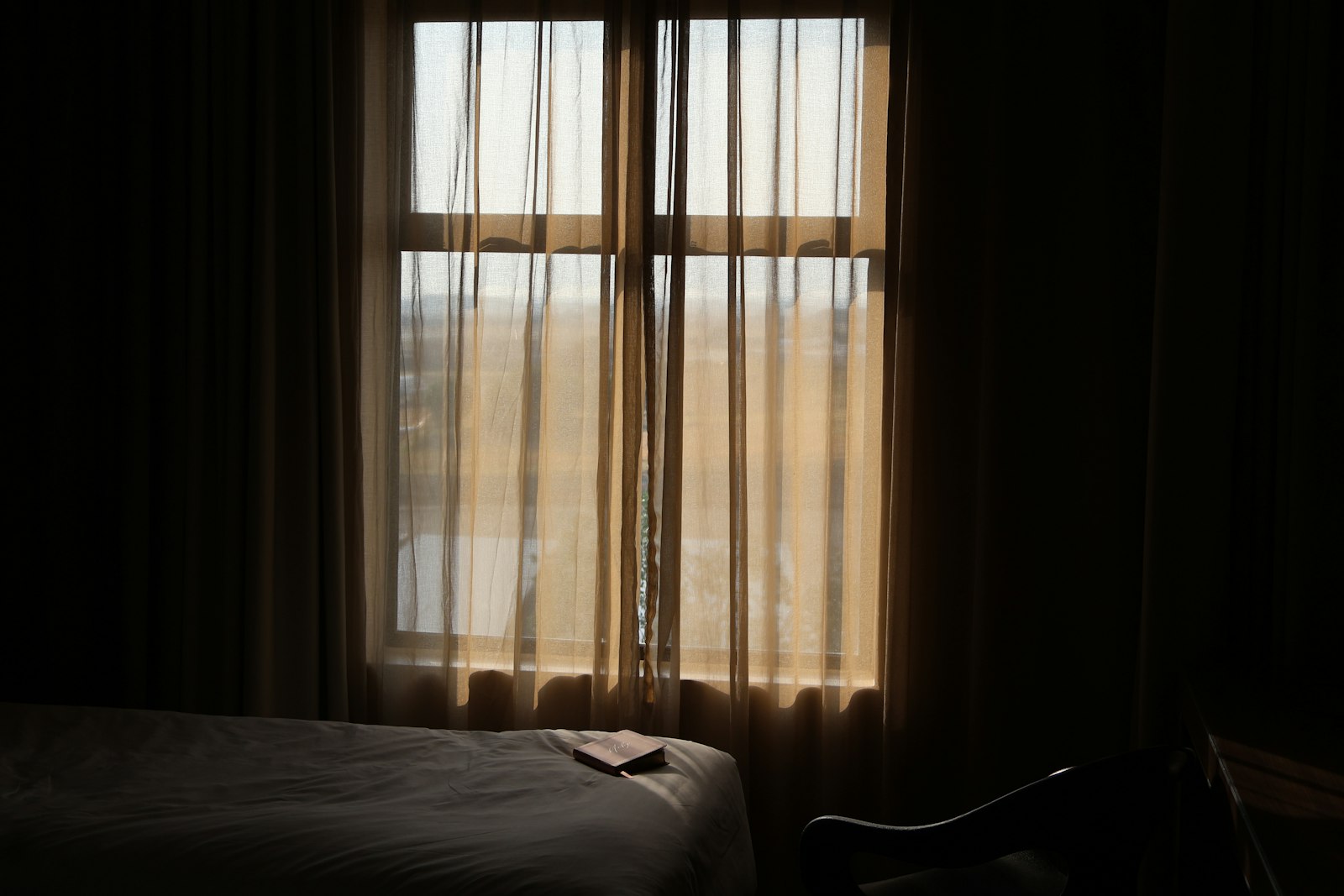 A dim room with sheer curtains at the window and a book left open on the bed